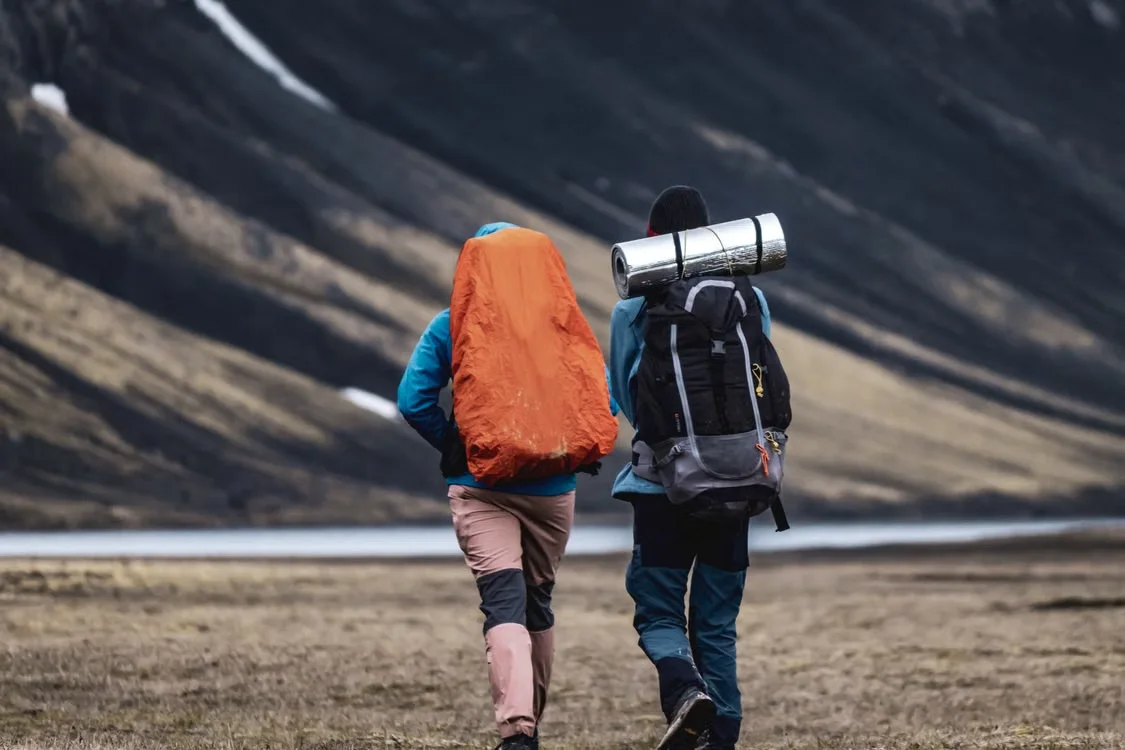 Two explorers trekking beside a mountain lake with backpacks and camping gear.