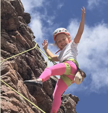 Aspen Owen in a climbing helmet and harness smiling while rock climbing on a rope against a blue sky.