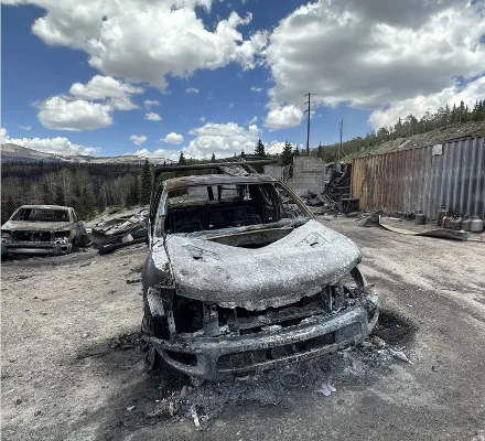 The burned front end of a pickup truck with other destroyed vehicles and fire debris behind it.